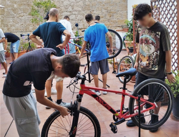 Dos jóvenes arreglan la rueda de una bicicleta mientras al fondo otros jóvenes siguen las indicaciones de los monitores