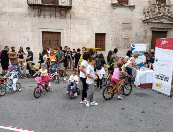 Grupo numeroso de adultos y niños en la calle, a pie y en bicicleta, junto a una mesa informativa