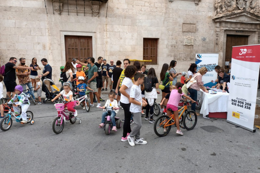 Grupo numeroso de adultos y niños en la calle, a pie y en bicicleta, junto a una mesa informativa